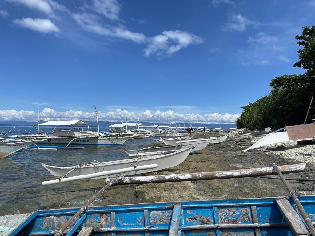 En rad vita bangkas förtöjda längs en klippig strand, små utriggarkanoter uppdragna på klipporna och en blå båt med flagnande färg i förgrunden, under en blå himmel med vita moln.