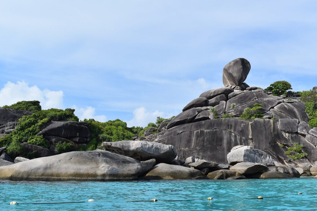 Det enorma balanserande granitblocket på ö 8 kröner en hög av släta grå klippor vid kanten av turkost vatten, under en disig blå himmel, med en rad bojar i förgrunden