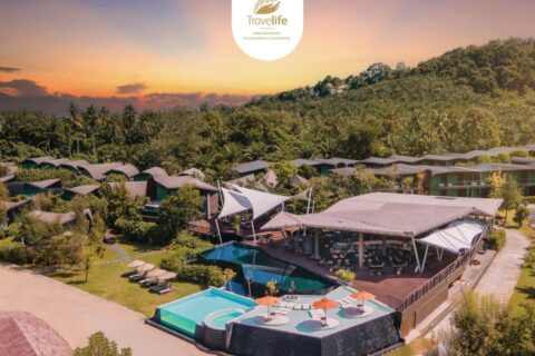 Flygbild över Kalima Resort Khao Lak i solnedgången, med pooler och svängda tak vid foten av en skogklädd kulle