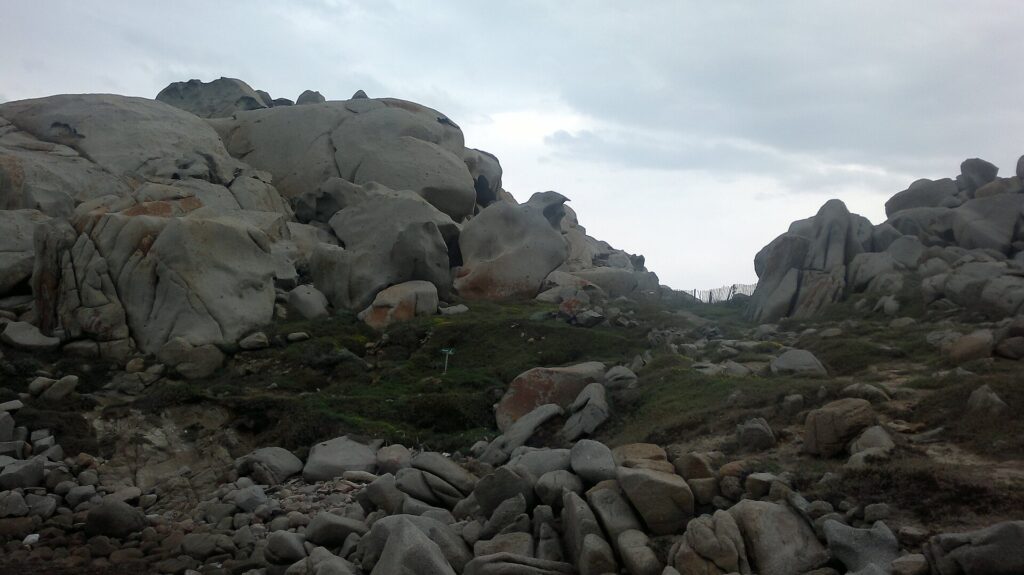 En samling stora, rundade grå granitblock urholkade av erosionen och fläckade av orange lavar, runt en kortklippt gräsmatta där en stig kantad av ett trästaket leder mot havet, under en mulen himmel.