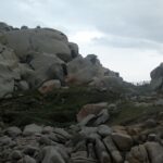 En samling stora, rundade grå granitblock urholkade av erosionen och fläckade av orange lavar, runt en kortklippt gräsmatta där en stig kantad av ett trästaket leder mot havet, under en mulen himmel.