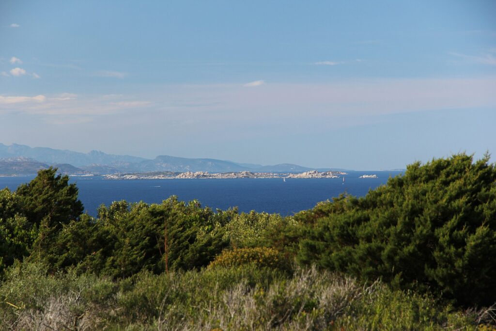 Vy från kustens macchia mot en lång, låg ö av ljusa granitblock i ett mörkblått hav, med en segelbåt vid dess fot och blåaktiga berg vid horisonten.
