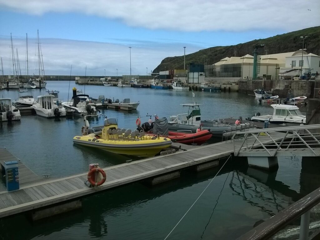 Ribbåtar, en gul med rader av säten och en röd, förtöjda vid bryggan i marinan i Vila do Porto, med segelbåtar och fiskebåtar bakom under en grön klippa och molnig himmel