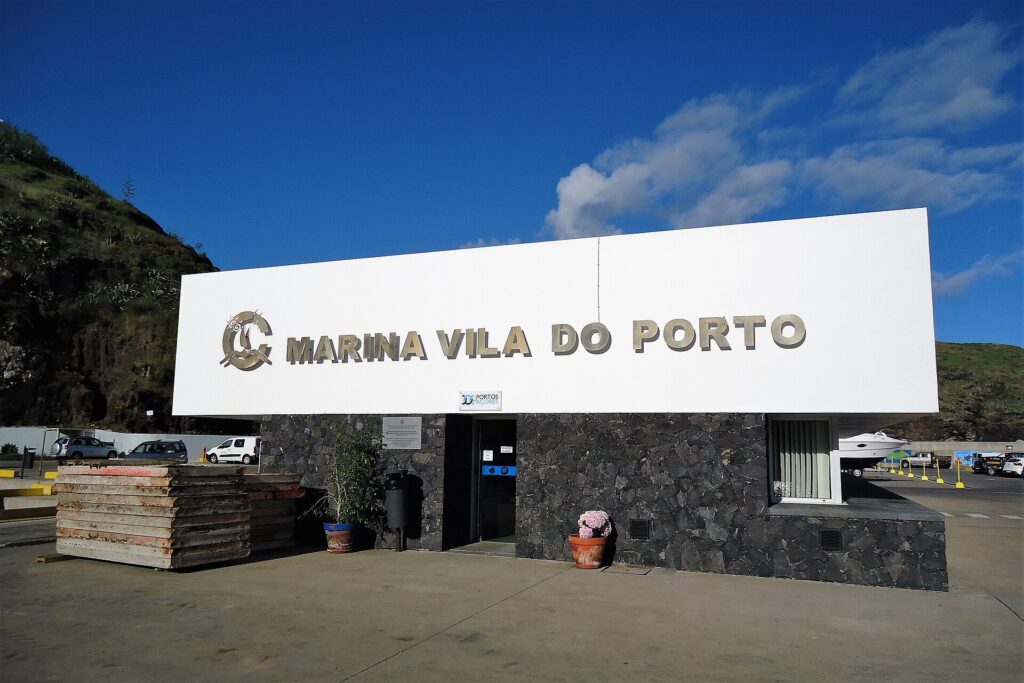 Marinans mottagningsbyggnad i Vila do Porto, en stor vit skylt med metallbokstäver på en svart basaltmur, under blå himmel, med en grön klippa till vänster