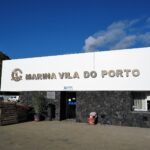 Marinans mottagningsbyggnad i Vila do Porto, en stor vit skylt med metallbokstäver på en svart basaltmur, under blå himmel, med en grön klippa till vänster