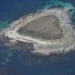 Flygbild av en liten holme av grå granithällar, i mitten täckt av torrt gräs där raserade stenmurar anas, kantad av en ljus strand och omgiven av turkosa grund och djupblått vatten.