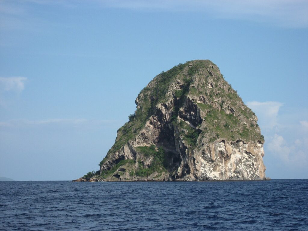 Rocher du Diamant sedd på nära håll från havet, en massa av grå klippa med grottor, vita strimmor och växtlighet på toppen, ovanför mörkblått hav