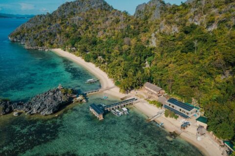 Flygbild över Sangat Island Dive Resort, med strand, brygga och båtar nedanför klipporna