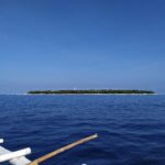 Den flata, skogklädda ön Balicasag vid horisonten, med sin vita fyr och sin rand av ljus sand, sedd från en bangka vars vita utriggare fyller förgrunden, under en molnfri blå himmel.