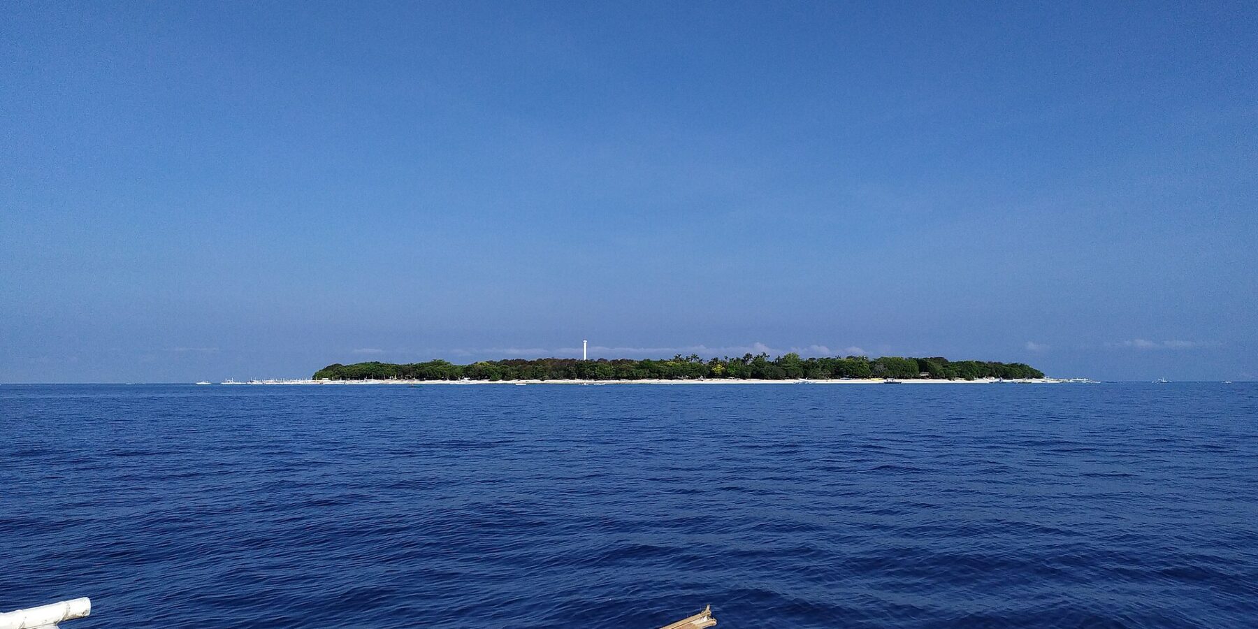 Den flata, skogklädda ön Balicasag vid horisonten, med sin vita fyr och sin rand av ljus sand, sedd från en bangka vars vita utriggare fyller förgrunden, under en molnfri blå himmel.
