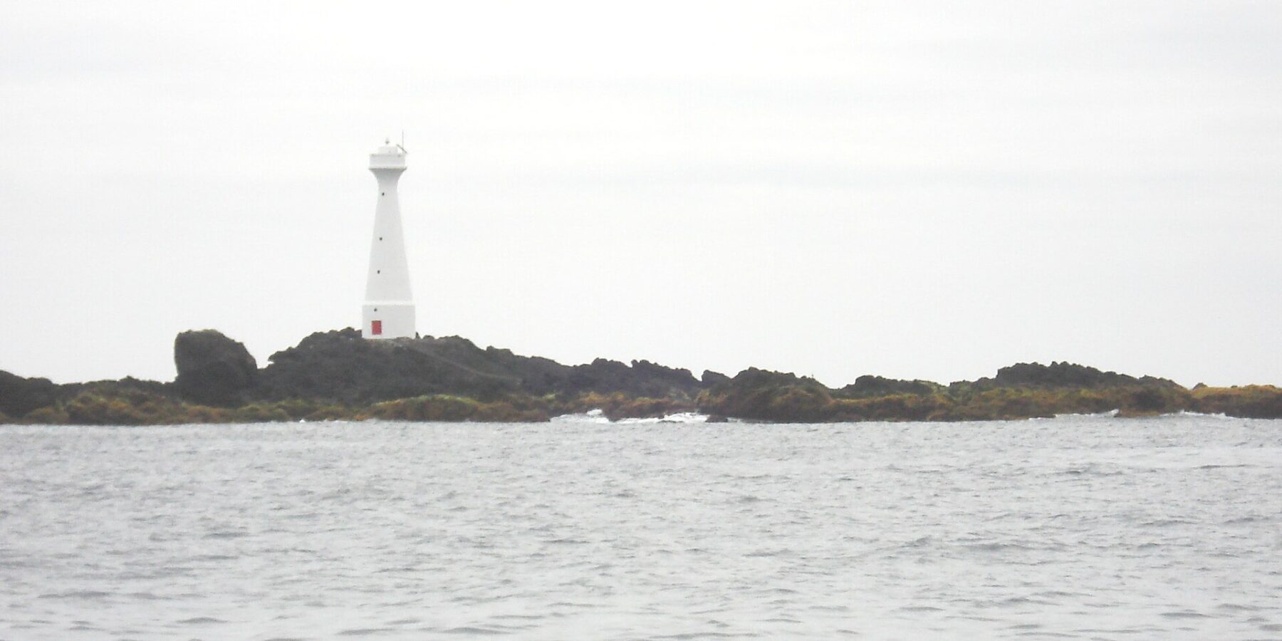 En smal vit fyr med röd dörr reser sig på den låga, klippiga holmen Formigão, med mörka klippor kantade av brunalger, ovanför ett grått och upprört hav under mulen himmel