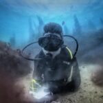 En dykare i svart dräkt, liggande på en sandbotten mellan två klippor täckta av bruna alger, skriver på en vit skrivtavla, med siluetter av tare mot det blå bakom sig.