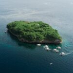 Flygbild av Pescador Island, en klippa täckt av grön växtlighet med en vit fyr, omgiven av ett tiotal dykbåtar av bancatyp på mörkblått hav