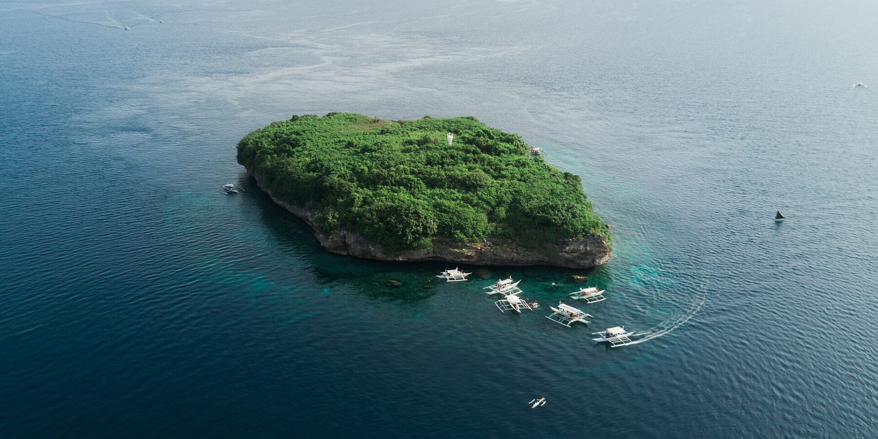 Flygbild av Pescador Island, en klippa täckt av grön växtlighet med en vit fyr, omgiven av ett tiotal dykbåtar av bancatyp på mörkblått hav