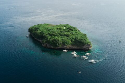 Flygbild av Pescador Island, en klippa täckt av grön växtlighet med en vit fyr, omgiven av ett tiotal dykbåtar av bancatyp på mörkblått hav