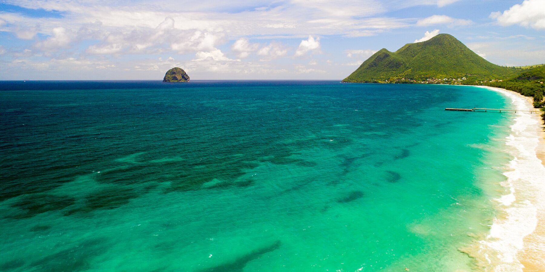 Flygbild av den långa stranden i Le Diamant kantad av turkost vatten, med Rocher du Diamant ute i havet och Morne Larcher, en grön kulle, till höger