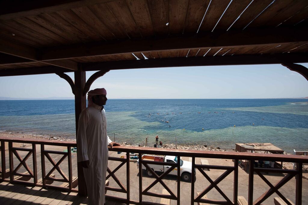 Från en träterrass ser en man i galabeya ut över bassängen i Blue Hole, kantad av ljust rev, där badare simmar