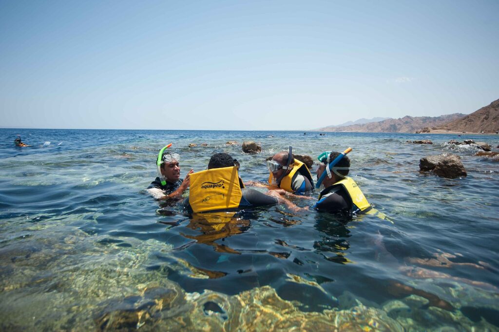 Simmare i gula västar, med mask och snorkel, i det klara vattnet i Blue Hole, med Sinais berg i bakgrunden